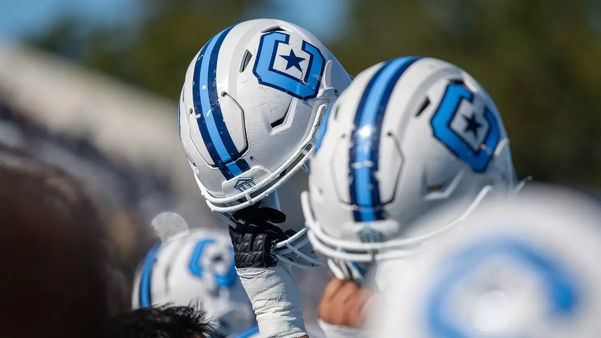 Athletes lift their white football helmets, featuring blue stripes and star-themed decals, in the air.