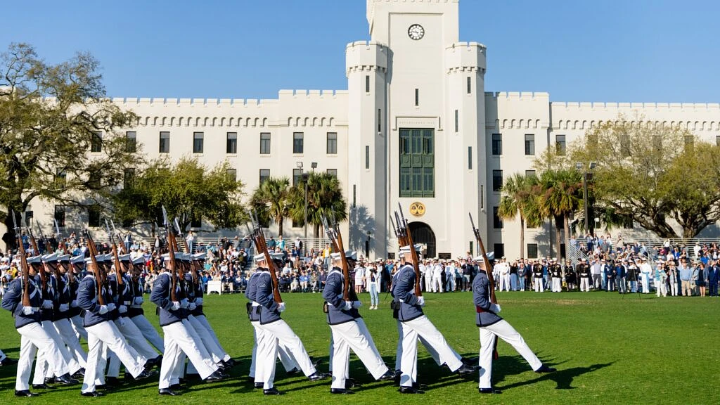 A group of cadets in uniform march in formation on a grassy field in front of a large, castle-like building, watched by a crowd.