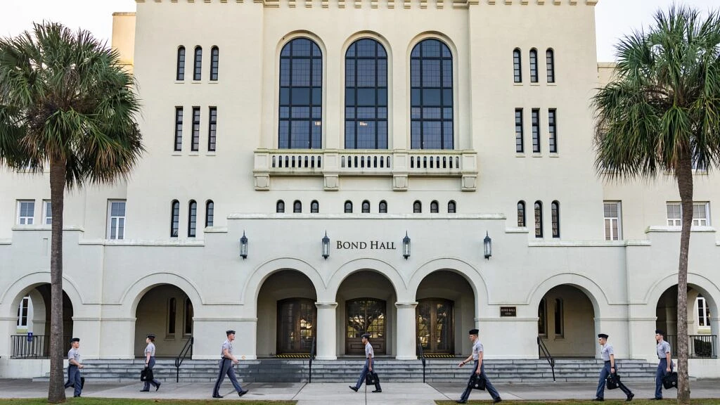 Several people dressed in uniform walk past an imposing building labeled "Bond Hall," flanked by palm trees.